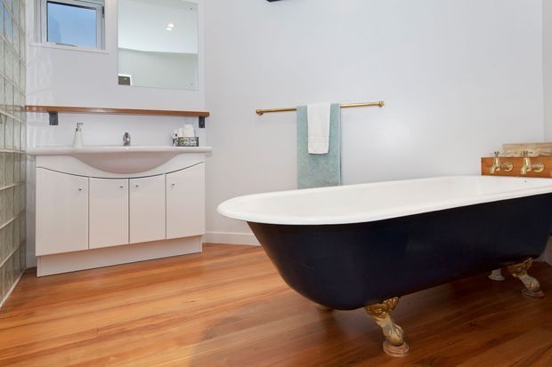 A bright, spacious bathroom featuring a freestanding black clawfoot bathtub with gold-toned feet and fixtures, a curved white vanity with integrated basin, timber countertop shelf, mirrored cabinet, glass-block wall, and warm timber flooring. Soft blue towels and brass/gold hardware add subtle colour accents.