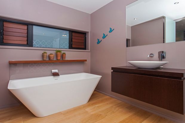 Contemporary bathroom featuring a freestanding white bathtub, wall-mounted dark timber vanity with a white vessel basin, large mirror, recessed ceiling lights, and a horizontal window with timber shutters. The room has mauve painted walls, warm timber flooring, and a simple floating timber shelf with small pots and decorative bird accents.