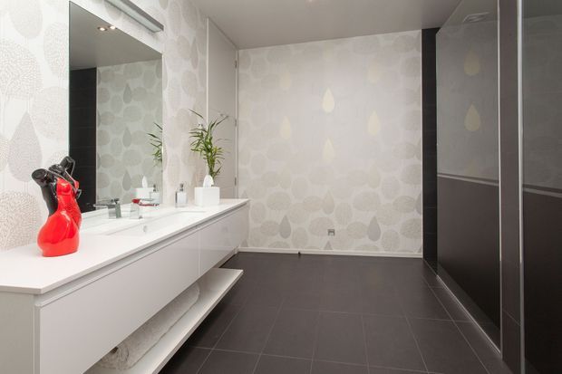 Contemporary bathroom with a long white floating vanity, integrated basin, large wall mirror, patterned pale wallpaper, dark charcoal floor tiles, and tall dark built-in cabinetry. A red decorative object and potted plant provide small accents.
