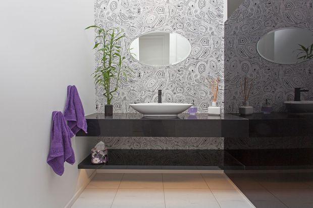 Contemporary bathroom vanity with a white vessel basin, wall-mounted matte dark cabinetry, large oval mirror, and an intricate grey patterned feature wall. Purple towels and a small plant add colour, while the mirror reflects the vanity and patterned wall.