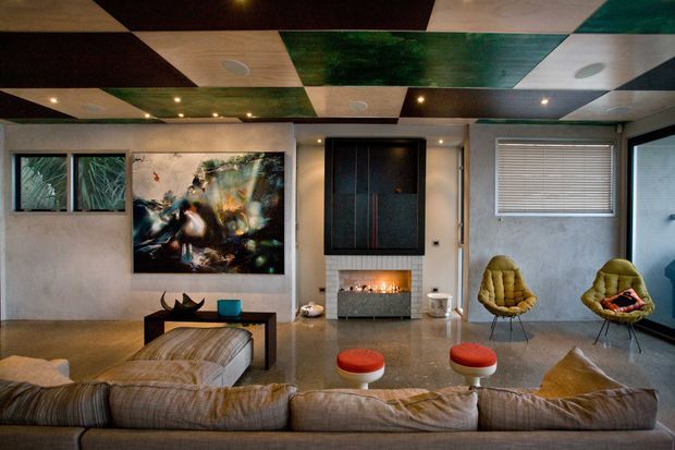 A stylised contemporary living room viewed from behind a large sectional sofa. The room features a polished concrete floor, textured pale walls, a dramatic geometric patchwork ceiling with timber, dark and green panels, recessed lighting, a central fireplace with built-in dark joinery, sculptural lounge chairs, and abstract artwork.