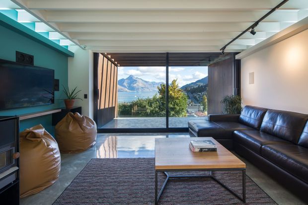 Bright contemporary living room with exposed white ceiling beams, a dark leather sectional sofa, beanbag seating, a low timber coffee table, and a large area rug. Full-height glazed sliding doors open to a scenic mountainous lakeside view, while a teal feature wall frames the television and built-in shelving.
