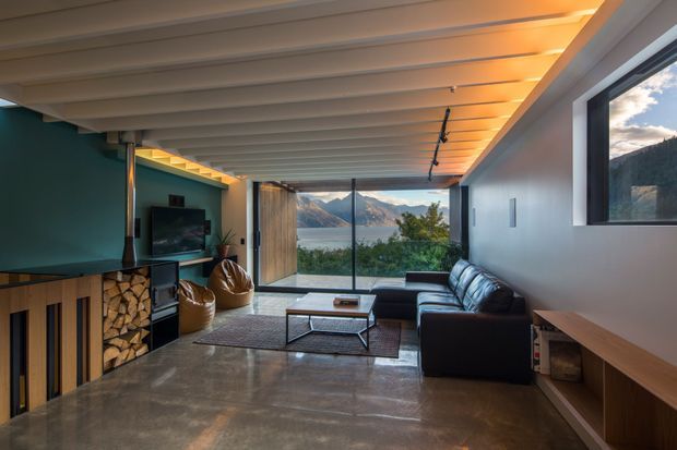 A contemporary living room with polished concrete flooring, a dark sectional sofa, woven lounge chairs, a low coffee table, and a built-in fireplace with exposed firewood storage. The room has a striking teal feature wall, warm timber-lined ceiling, concealed lighting, and expansive glazing opening to a mountain and lake view.