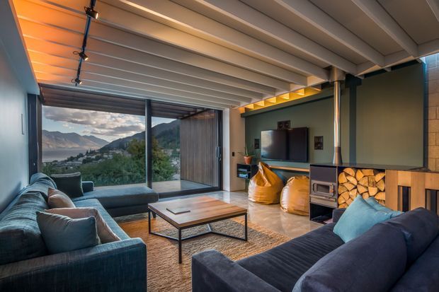 A warm, modern living room with exposed white-painted ceiling beams, dark walls, timber flooring, and large sliding glass doors framing a mountain and lake view. The room includes two deep blue sofas, a black-framed coffee table, a wall-mounted television, a compact black fireplace with stacked firewood, and warm integrated lighting.