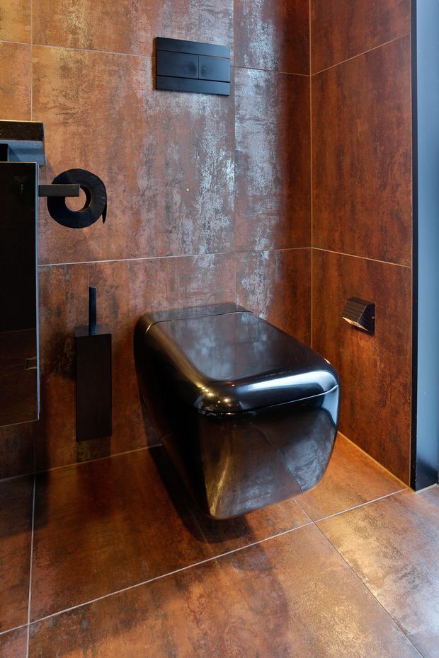 A compact, dramatically styled bathroom featuring a wall-hung black toilet with matching black flush plate and wall-mounted accessories. Large rust-brown, weathered metallic-look tiles cover the walls and floor, creating a dark industrial atmosphere. Black fixtures and a reflective metallic toilet contrast with the warm textured tile surfaces.