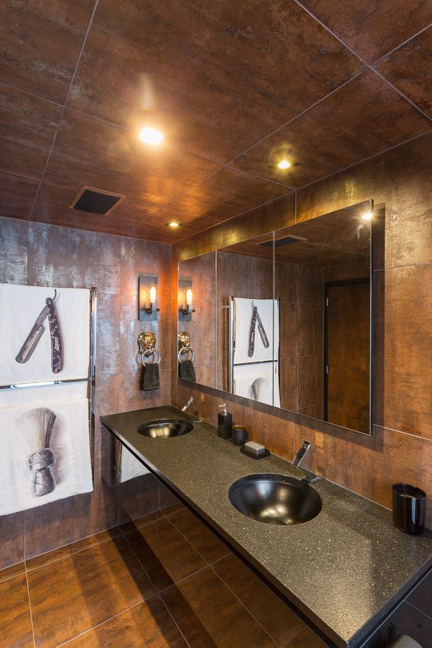 A compact, dramatic bathroom with two black vessel basins set into a dark speckled countertop, bronze-brown metallic-look tiled walls and ceiling, matching dark floor tiles, a large wall mirror, black fixtures, and warm recessed and wall lighting. Decorative black-and-white towels add contrast.
