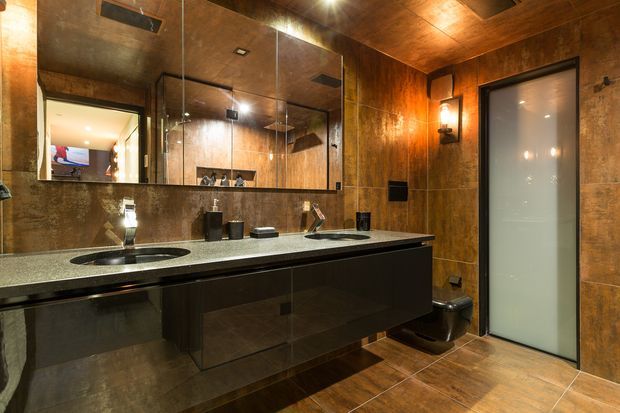 A sophisticated double-vanity bathroom with dark glossy cabinetry, a long light stone benchtop, integrated basins, and wall-to-wall mirror panels. Warm brown tiled or stone-look wall and floor surfaces create a richly textured, spa-like atmosphere. A frosted-glass door is visible on the right, with contemporary wall lighting and dark fixtures.