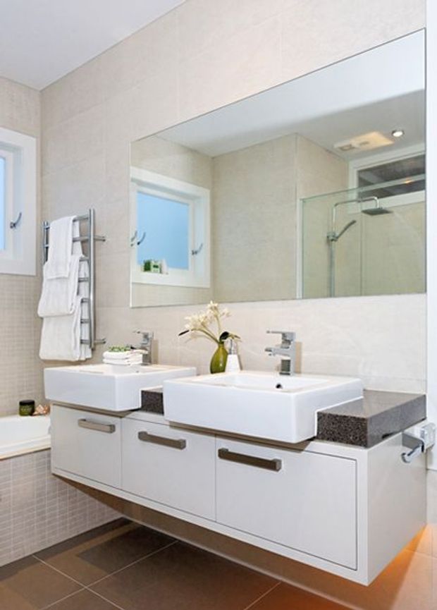 Bright contemporary bathroom featuring a floating double vanity with two white vessel basins, grey stone benchtop, wall-to-wall mirror, glass shower enclosure, wall-mounted towel warmer, and tiled walls and floor.