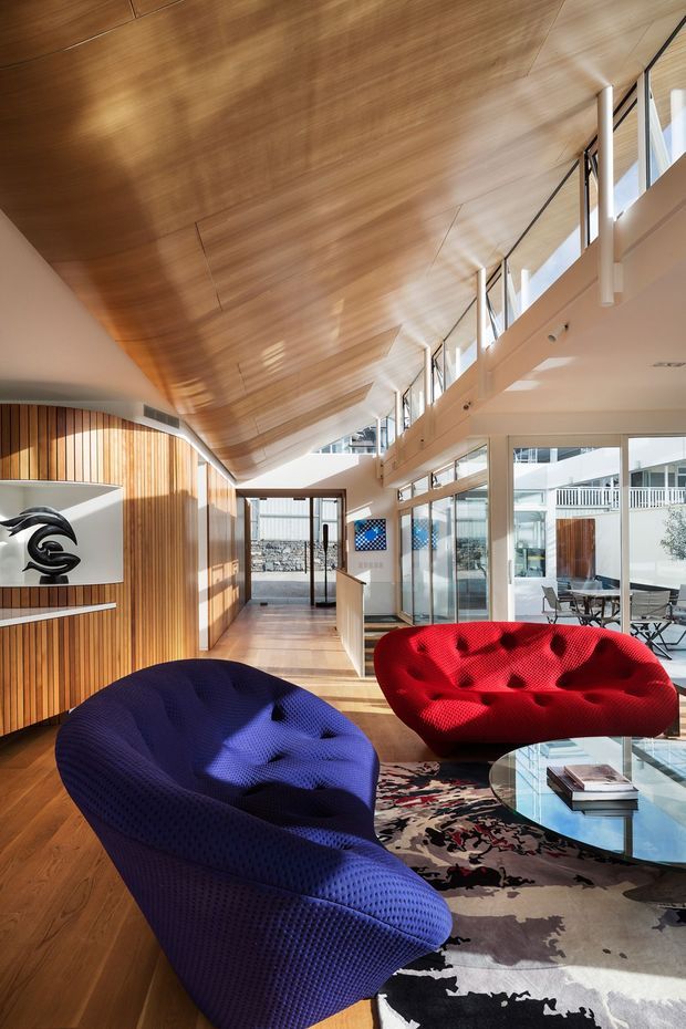 A bright, double-height contemporary interior lounge with a dramatic sloped timber-lined ceiling, extensive clerestory glazing, and a long corridor-like layout. Sculptural deep-blue and red lounge chairs surround a round glass coffee table on a bold patterned rug. Vertical timber wall panelling, white structural elements, and views into a courtyard or adjacent glazed area create a refined, gallery-like atmosphere.
