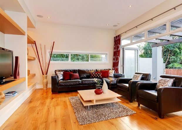 Bright contemporary living room with warm timber flooring, built-in white shelving and television joinery, a black leather sectional and armchairs, layered coffee tables, and a textured area rug. Large clerestory and full-height glazing bring in abundant daylight, with red patterned curtains adding a strong accent.