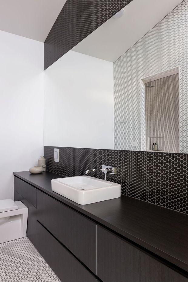 A contemporary bathroom featuring a long dark timber-look floating vanity with integrated drawers, a white vessel basin, and a wall-mounted chrome tap. Black hexagonal mosaic tiles form the splashback and continue across the ceiling feature, while small white hexagonal tiles cover the floor and adjacent wall. A large frameless mirror reflects the tiled wall and shower area. A white toilet is partially visible at left.