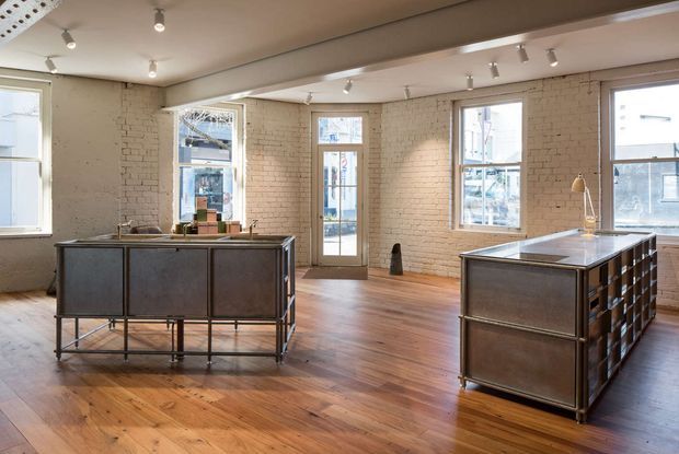 Open-plan loft-style interior with exposed painted brick walls, warm timber flooring, large industrial windows, and two substantial dark metal kitchen islands or workbenches. Track lighting and exposed structural detailing create an industrial studio atmosphere, with urban buildings visible through the glazing.