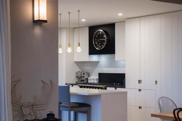 A refined contemporary kitchen with a compact island and bar seating, extensive floor-to-ceiling white built-in cabinetry, integrated appliances, pale stone or light benchtops, and a dark feature panel displaying a round wall clock. Brass-toned pendant lights and discreet recessed ceiling lighting add warmth. Part of a dining area with a transparent chair and timber table is visible in the foreground.