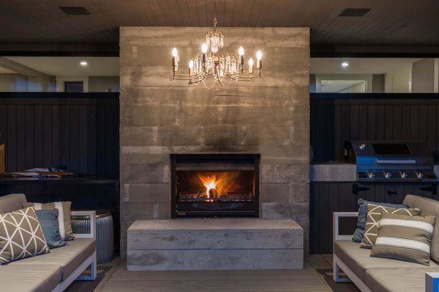 A moody, refined indoor entertaining and lounge area centred on a concrete fireplace feature wall with an active fire and a large crystal chandelier. Dark built-in cabinetry and integrated barbecue-style appliances flank the fireplace, while upholstered lounge seating sits in the foreground. The space has a timber-lined ceiling, dark timber joinery, and a warm, symmetrical composition.