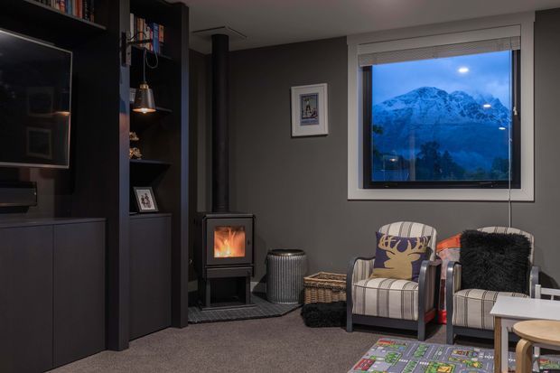 A compact, moody living room with dark built-in shelving and media cabinetry, a freestanding wood-burning stove, two upholstered armchairs, and a large window overlooking snow-covered mountains. The room has grey walls, dark joinery, carpeted flooring, layered lighting, and a cosy alpine atmosphere.