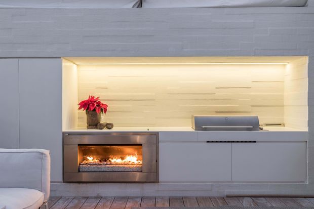 A built-in contemporary entertainment and fireplace wall in a light grey room. The feature unit has flush grey cabinetry, a recessed illuminated shelf with a textured light timber or painted finish, a linear gas fireplace, and a low-profile media component. Pale timber flooring and part of a light upholstered sofa are visible.