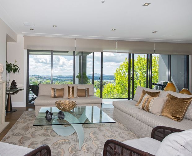 Bright, spacious living room with floor-to-ceiling glazing overlooking a leafy landscape and distant hills. The room features large light-upholstered sofas, patterned cushions, a sculptural glass coffee table, a large patterned rug, recessed ceiling lights, roller blinds, and dark-framed windows and doors.