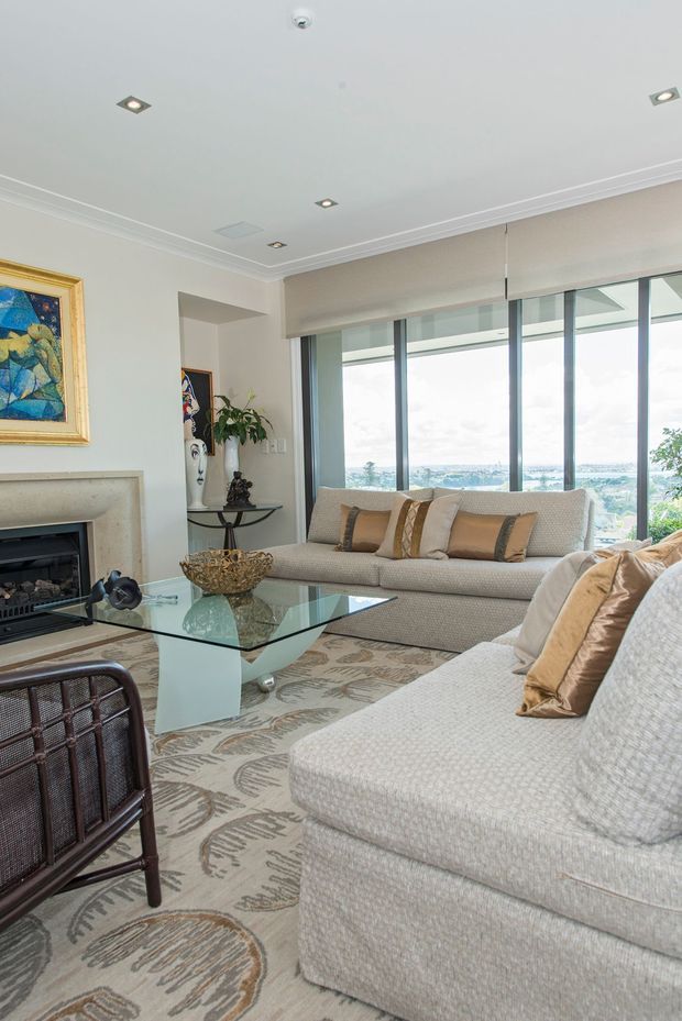 A bright, refined living room with large floor-to-ceiling windows overlooking a distant urban or coastal landscape. The room features two pale upholstered sofas with beige and tan cushions, a glass coffee table, patterned area rug, fireplace with a light stone surround, recessed ceiling lights, crown moulding, and a decorative framed artwork. Neutral finishes and abundant glazing create an elegant, airy atmosphere.