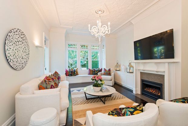 Bright, refined living room with white upholstered sofas and armchairs arranged around a glass coffee table and patterned rug. The room features a decorative white fireplace with an inset fire, built-in cabinetry, bay-style windows with a window seat, ornate ceiling mouldings, a textured ceiling, and a white chandelier. Colourful patterned cushions and chair upholstery provide vivid accents against the predominantly white interior.