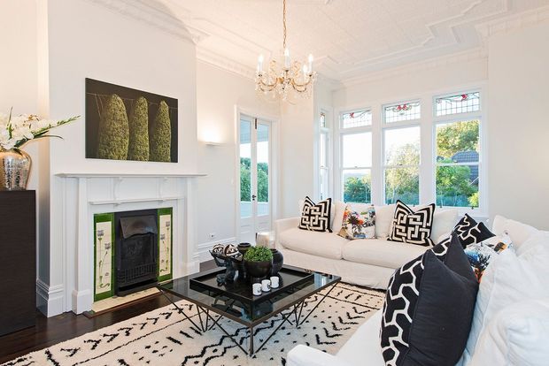 Bright, refined living room in a period-style home, featuring ornate ceiling detailing, decorative cornices, a crystal chandelier, white walls and fireplace surround, dark timber flooring, tall leadlight-style windows, and a large white sectional sofa. A black glass coffee table and monochrome geometric rug and cushions add contemporary contrast.