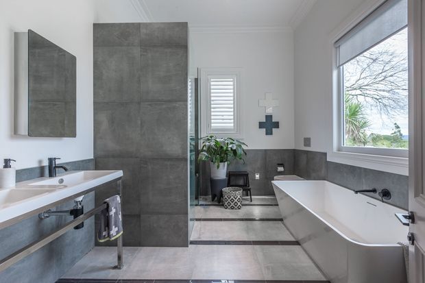 Bright contemporary bathroom with a freestanding white bathtub positioned beneath a large window, a wall-mounted vanity with exposed dark metal plumbing, and a walk-in shower enclosed by large grey stone-look wall panels. The room has pale tiled flooring with dark linear border accents, white walls with decorative ceiling moulding, plantation shutters, and restrained black and grey fixtures. A small plant and stool add natural detail.
