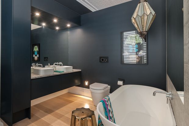 A stylish dark-toned bathroom with deep charcoal walls, a double vanity with white vessel basins, a freestanding white bathtub, and a modern toilet. Warm timber-look flooring contrasts with the dark surfaces, while a faceted pendant light, recessed spotlights, louvred window, and subtle under-vanity lighting add character and ambience.