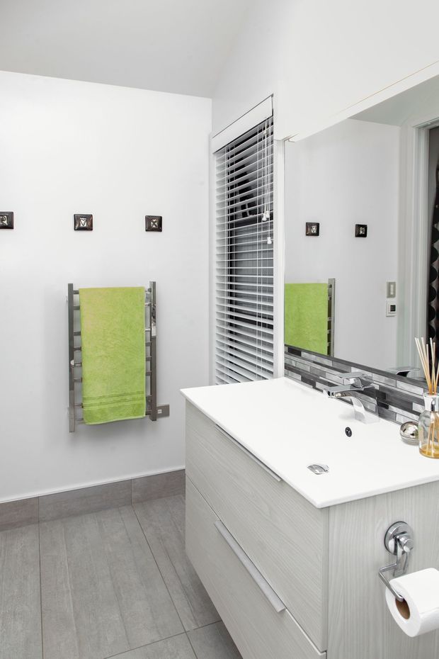 Bright contemporary bathroom featuring a wall-mounted light wood vanity with a white benchtop, integrated basin and chrome tapware. Large grey floor tiles, a mirrored wall, white plantation shutters, a heated towel rail with a lime-green towel, and grey mosaic wall tiling create a clean, modern finish.