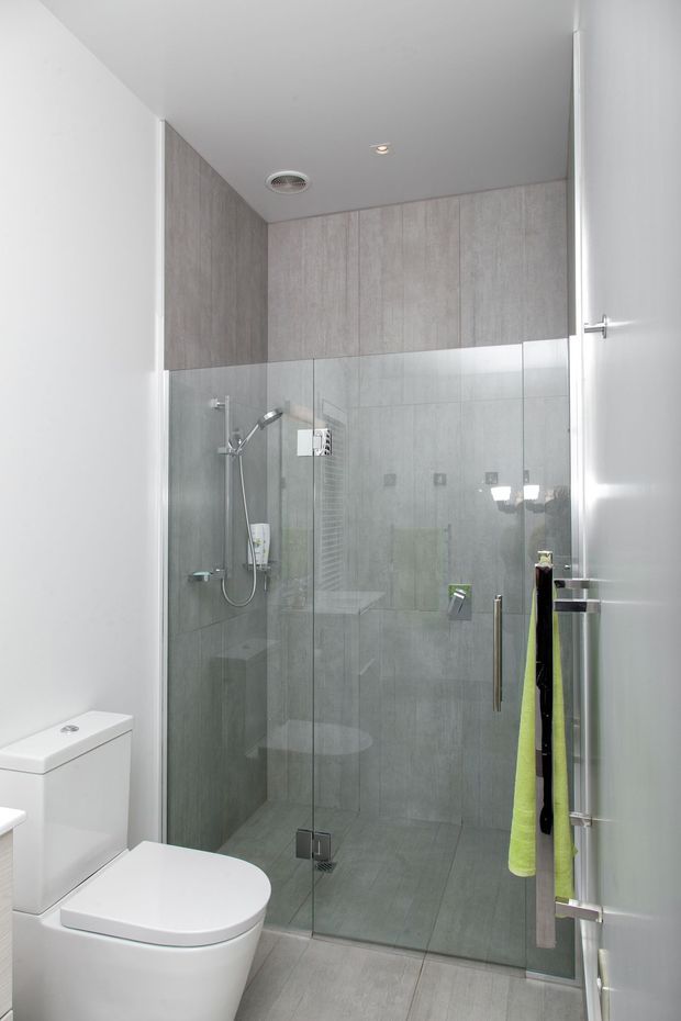 Compact contemporary bathroom featuring a walk-in glass shower enclosure with grey tiled walls and floor, a wall-mounted shower system, white toilet, and light wood-look wall panels above the shower. The space has a clean, bright, minimalist fit-out with chrome hardware and recessed ceiling lighting.