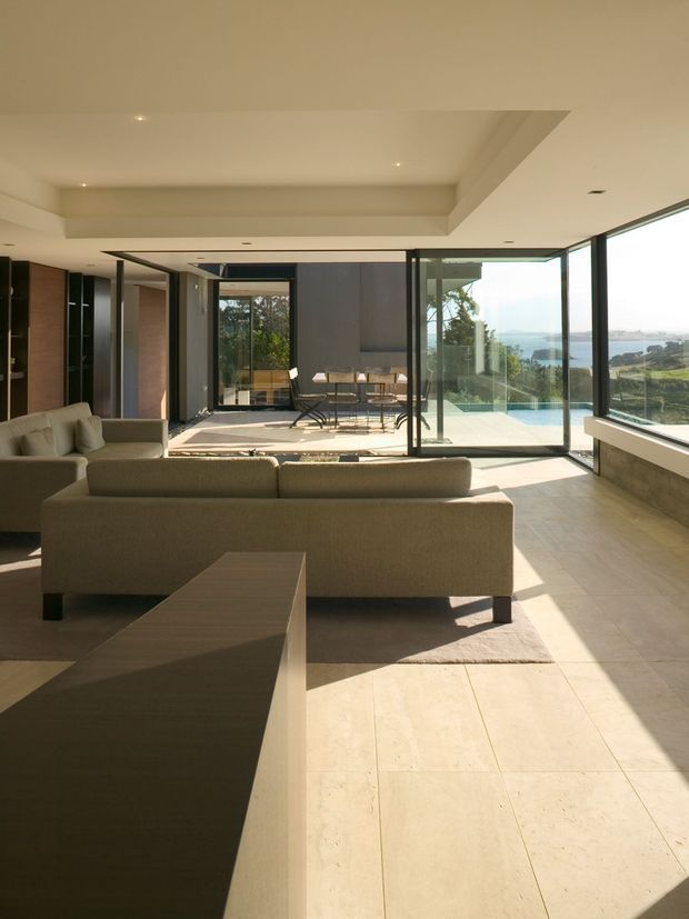 A bright, open-plan contemporary living room opening onto a terrace and swimming pool through extensive floor-to-ceiling glass doors. The room features a large neutral sectional sofa, a low angular coffee table, pale large-format tiled flooring, dark built-in cabinetry, a recessed tray ceiling with downlights, and a dining area beyond. Coastal scenery is visible through the glazing.