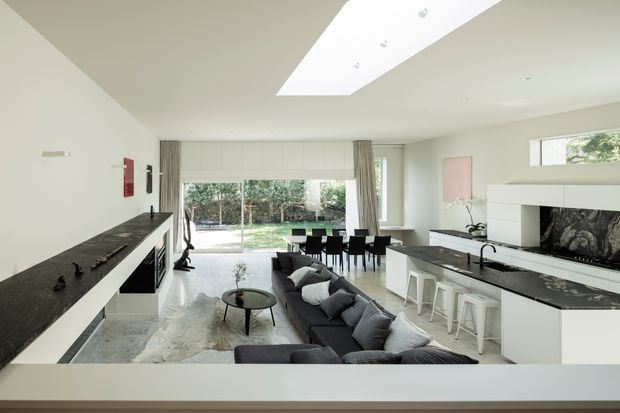 Bright open-plan contemporary interior combining a kitchen, dining area and living room. White walls and cabinetry contrast with dark stone benchtops, while a long charcoal sectional faces a sleek fireplace with a dark stone feature surround. Large glazed doors open to a garden, and a linear skylight brings additional daylight into the space.