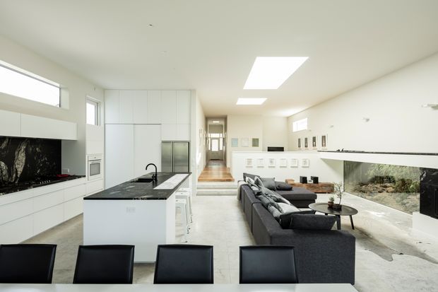 Bright open-plan kitchen and living room with a minimalist white fit-out. The kitchen has extensive white built-in cabinetry, a dark stone island with sink, integrated appliances, and a black marble-look splashback. A dark sectional sofa faces a low TV console, while a large internal glazed feature wall reveals a planted rock garden. Skylights and clerestory windows provide abundant natural light, and a timber-floored hallway extends beyond the living area.