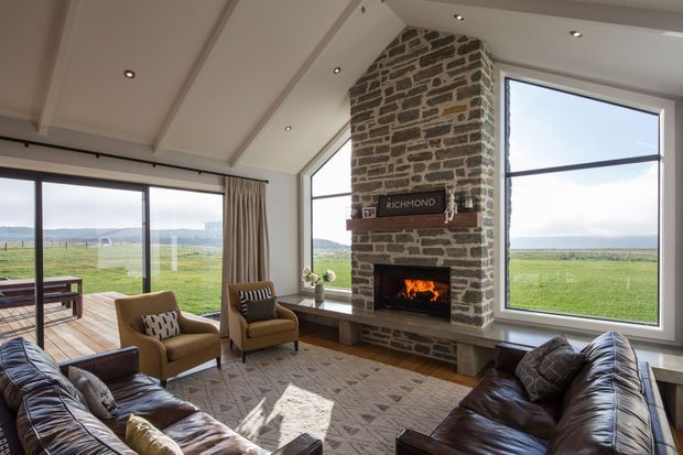 A spacious living room with expansive floor-to-ceiling windows overlooking open green fields and distant water or coastline. The room features a large rough-stone fireplace chimney with a built-in wood-burning fire, a projecting stone hearth, exposed timber ceiling beams, leather sofas, upholstered armchairs, a patterned area rug, and timber decking visible through the glazing. The interior has a warm, rustic lodge-like character with abundant natural light.