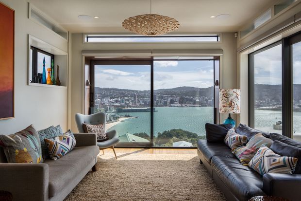 Bright contemporary living room with expansive floor-to-ceiling glazing overlooking a turquoise harbour and dense hillside cityscape. The room features two sofas, a lounge chair, patterned cushions, a large textured rug, built-in display shelving, a sculptural woven pendant light, and neutral warm-toned finishes.