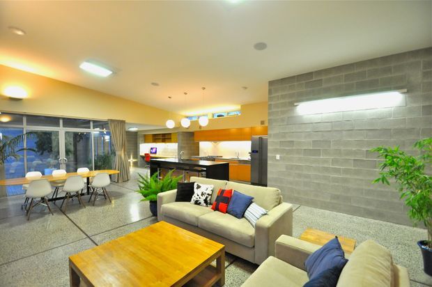 Open-plan contemporary living and dining area with a kitchen in the background. The space features polished concrete or terrazzo-style flooring, a grey block feature wall, warm orange-toned kitchen cabinetry, expansive glazing with curtains, and neutral sofas around a timber coffee table. Recessed and linear lighting creates a warm, inviting atmosphere.
