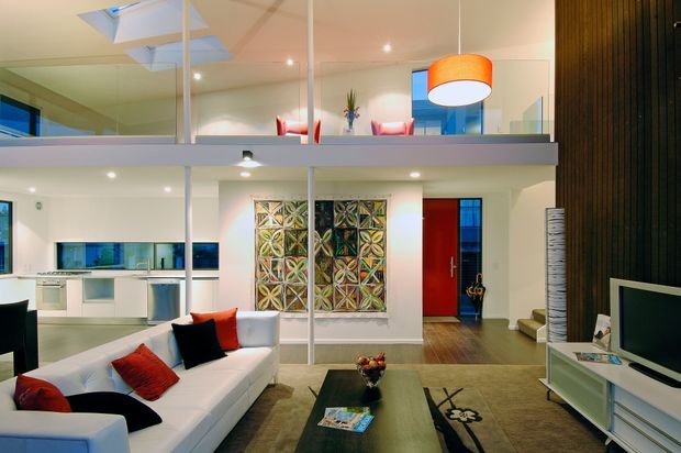 Open-plan contemporary living room with a white sectional sofa, dark coffee table, television console, and decorative feature artwork. A compact white kitchen is visible to the left, while a mezzanine-level space with glass balustrades overlooks the room. The interior has recessed lighting, a bright orange pendant, a bold red door, polished timber flooring, and a dark vertical timber feature wall.