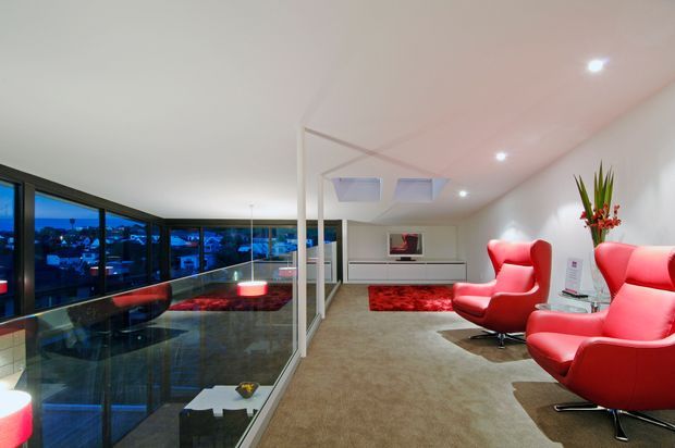A contemporary open-plan living area overlooking a city at dusk through extensive floor-to-ceiling glazing. The room has a neutral carpeted floor, white walls and ceiling, a sleek low built-in media cabinet, a red area rug, and several bold red sculptural lounge chairs. A glass balustrade separates the living space from a lower level, while recessed lighting and cooling vents create a clean modern interior.