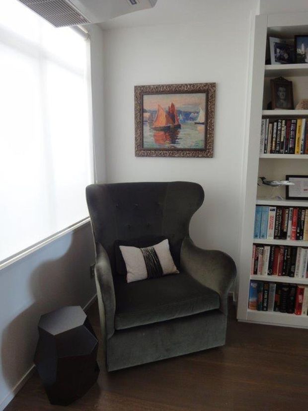 A compact, softly lit reading nook featuring a dark charcoal wingback armchair with a small accent cushion, a faceted black side table, dark timber flooring, white walls, and a full-height built-in bookshelf filled with books. A framed maritime painting hangs above the chair, while a large bright window with a translucent blind provides natural light.