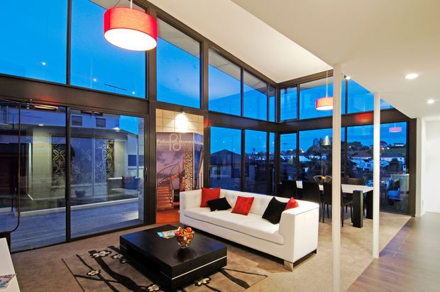 A contemporary open-plan living room with expansive floor-to-ceiling glazing overlooking an urban landscape at dusk. The space features a white sofa with red and black cushions, a low black coffee table, a patterned rug, pendant lighting, dark-framed glass walls and doors, and a visible dining area beyond. The high glazing and blue evening light create a polished, dramatic atmosphere.