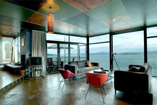 A mid-century-inspired living room with expansive floor-to-ceiling glazing overlooking a calm bay and distant hills. The room features a polished concrete floor, dark-framed glass doors and windows, a patterned tiled ceiling, sculptural pendant light, large sofas, and colourful retro lounge chairs arranged around a low coffee table.
