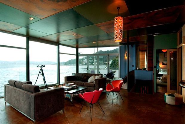 A dramatic waterfront living room with full-height corner glazing overlooking the water. The room features dark brown polished concrete flooring, a richly patinated metallic ceiling, exposed dark timber beams, a large sectional sofa, sculptural red lounge chairs, a coffee table, and warm pendant lighting. A hallway or adjoining room is visible at right.