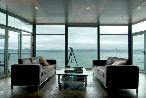 A contemporary waterfront living room enclosed by expansive floor-to-ceiling glazing, with two facing sofas, a low rectangular coffee table, and a tripod near the window. The room has a dark polished floor, a reflective panelled ceiling with recessed spotlights, dark window and door frames, and uninterrupted views across the water and distant hills.