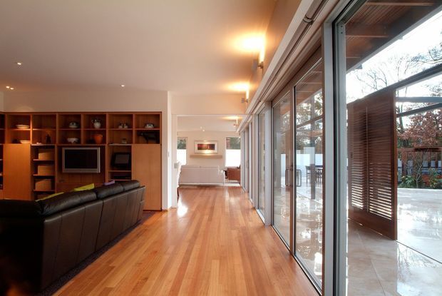 A warm, open-plan living room with built-in timber entertainment shelving and cabinetry, a wall-mounted television, and a dark leather sectional sofa. Polished timber flooring extends toward a bright glazed edge with large sliding glass doors opening to a covered outdoor terrace. Recessed and wall-mounted lighting create a welcoming atmosphere, while timber screening is visible outside.