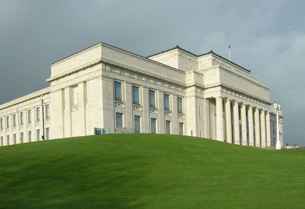 Exterior view of a large neoclassical civic or institutional building set on a broad manicured grassy slope. The pale stone facade features symmetrical wings, tall rectangular windows, strong horizontal cornices, decorative classical detailing, and a prominent colonnaded entrance with tall columns. The image is photographed under a cloudy sky.