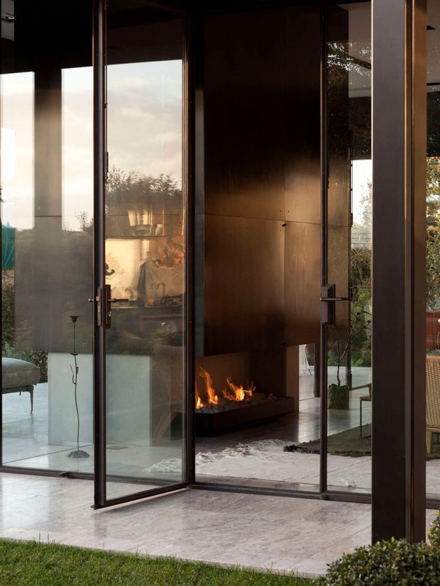 A dark, refined interior viewed through large open glazed doors, featuring a linear fireplace set within a tall bronze-brown wall panel, polished concrete flooring, and strong indoor-outdoor connection. Reflections and glazing reveal a softly furnished living area, while warm firelight contrasts with daylight and garden views.