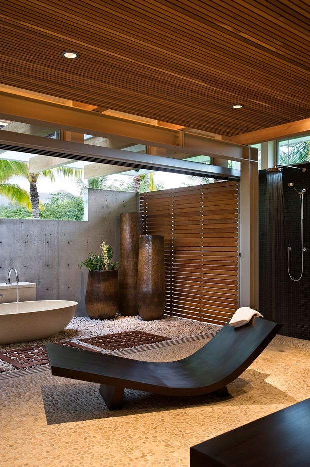 A luxurious open-air bathroom with a freestanding oval bathtub, wall-mounted shower, timber slatted privacy screen, concrete walls, and a sculptural dark wood reclining bench. The space features a richly slatted timber ceiling with recessed lighting, pebble mosaic flooring, decorative river stones, planted vessels, and tropical greenery visible beyond the openings.