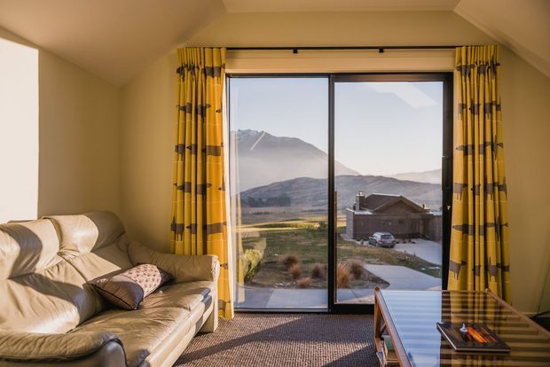 A warm, compact living room with a tan leather sofa, patterned yellow-and-dark curtains, carpeted floor, and a large sliding glass door offering expansive mountain and rural valley views. A low coffee table and media or storage unit sit in the foreground, while the sloped ceiling creates a cozy upper-level feel.