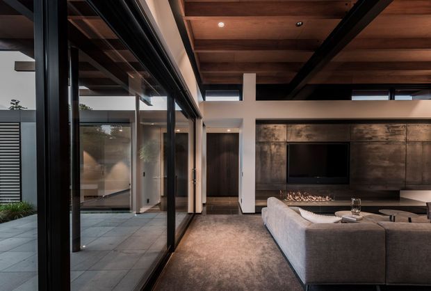A sophisticated contemporary living room viewed from inside, featuring a large charcoal sectional sofa, expansive black-framed floor-to-ceiling glazing, a dark timber-lined ceiling, and a dramatic concrete or stone media wall with built-in fireplace and television. The space opens onto a paved outdoor courtyard, creating a strong indoor-outdoor connection. The palette is dark, warm, and refined with layered timber, concrete, glass, and neutral upholstery.