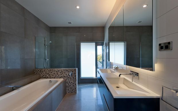 Contemporary bathroom with a long double vanity and integrated white basins beneath a large mirrored wall, a freestanding bathtub at left, and a separate glass-enclosed shower behind a low mosaic-tiled partition. Large-format grey wall and floor tiles create a sleek, restrained finish, with a frosted-glass door or window bringing in natural light.