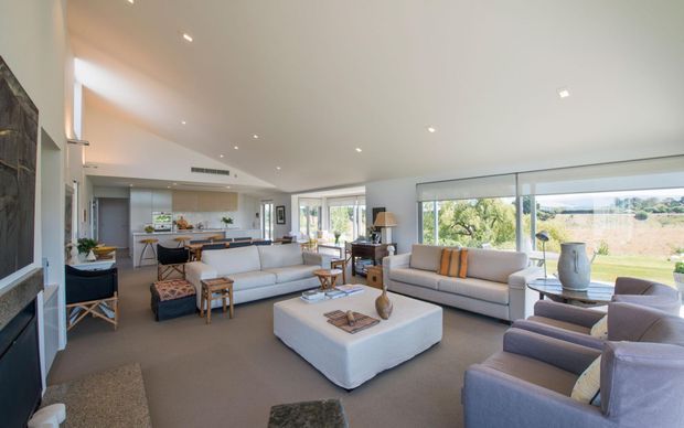 Bright, spacious open-plan living area with a contemporary neutral interior. The room features large floor-to-ceiling windows overlooking a landscaped outdoor setting, two pale sofas, blue-grey armchairs, a large upholstered square ottoman coffee table, recessed ceiling lighting, and an open kitchen and dining area beyond. A fireplace or dark feature wall is visible at the left edge, with timber cabinetry and subtle warm accents throughout.