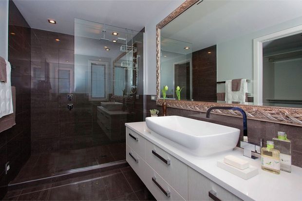 Contemporary bathroom with a large white vanity, above-counter vessel basin, dark brown tiled walls and floor, frameless glass shower enclosure, and an ornate framed mirror above the basin. The room has recessed ceiling lighting and a clean, moderately luxurious finish.